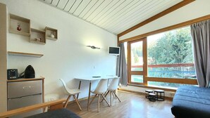 Een kleine kamer met een bed, een bureau, twee stoelen, een televisie en een raam met uitzicht op groen.