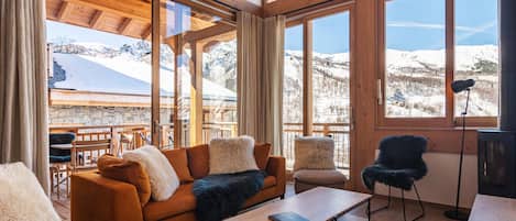 A cozy living room with a wooden ceiling, large windows with a snowy mountain view, a sofa, a coffee table, and a chair.