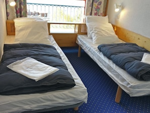 Two single beds with wooden headboards and white bedding, set against a blue polka dot carpet and a window with a metal railing offering an outdoor view.