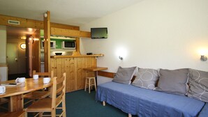 A room with a wooden dining table, chairs, a blue sofa, a small kitchen area with a microwave, and a television mounted on the wall.