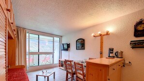 Una habitación acogedora con una cama de madera, una mesita con vasos, una mesa de comedor con sillas, un televisor montado en la pared y un reloj.