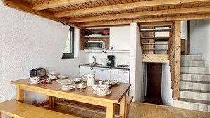 Puinen ruokapöytä on katettu lautasilla, kupeilla ja siinä on myös vedenkeitin. Taustalla näkyy keittiö, jossa on mikroaaltouuni, kahvinkeitin ja astianpesukone.