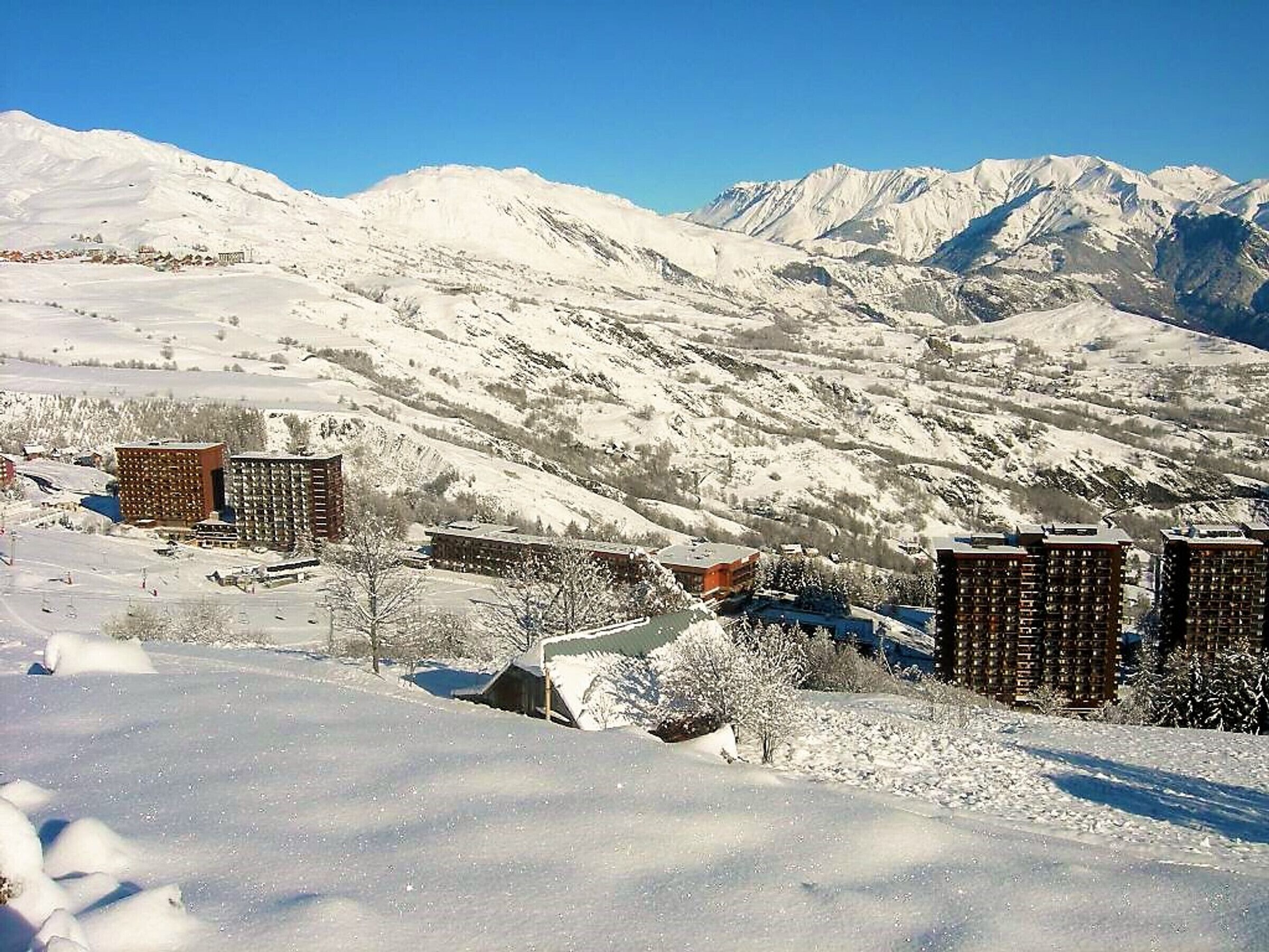 Eine verschneite Berglandschaft mit mehreren Gebäuden, darunter ein mehrstöckiges Hotel und ein Skiort im Hintergrund.