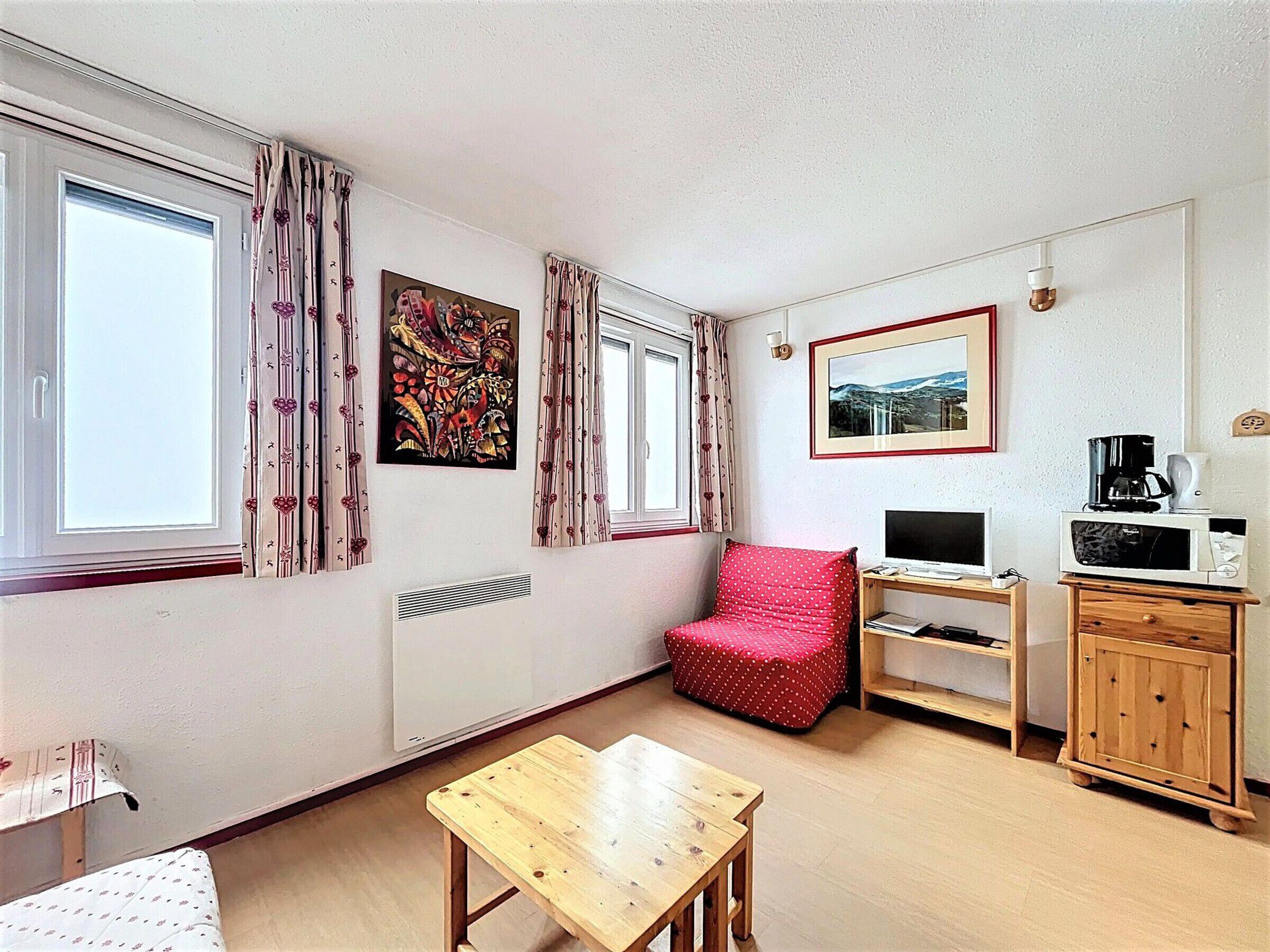 Una habitación pequeña y bien iluminada con una mesa de madera, una silla roja, un televisor, una cafetera y un cuadro en la pared.