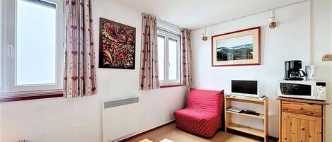 Una habitación pequeña y bien iluminada con una mesa de madera, una silla roja, un televisor, una cafetera y un cuadro en la pared.
