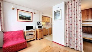 Una habitación compacta con un sofá rojo de lunares, una mesa de madera, una kitchenette con armarios de madera y una cama con cortinas a rayas.