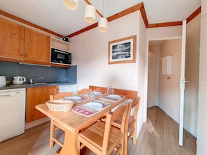 A compact kitchen with wooden cabinets, a microwave, and a small refrigerator. A dining table with chairs and a placemat is set up in the center. - Apartment 4 ppl near slopes with balcony (Les Menuires)