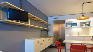 Een moderne keuken met witte kasten, een ingebouwde magnetron en een televisie op een plank. Er staan twee rode stoelen en een klein houten kruk.