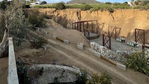 Ein Feldweg führt zu einer Holzterrasse mit Stühlen und Tischen, umgeben von einem angelegten Garten mit verschiedenen Pflanzen und einer Steinsäule.