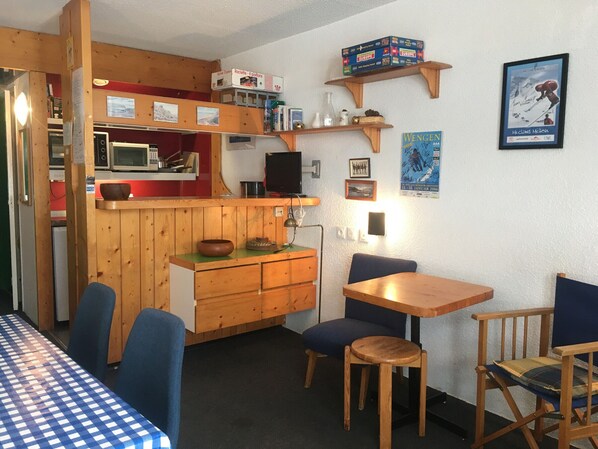 Een gezellige kamer met een houten tafel en stoelen, een kitchenette, een televisie en diverse decoraties aan de muren.