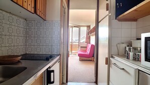 A compact kitchen with a sink, stove, and microwave. A pink chair is visible in the adjacent room.