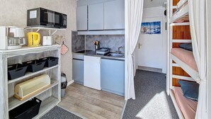 Cozinha compacta com micro-ondas, chaleira e pia. Existe uma pequena área de refeições com mesa e cadeiras. Uma cama beliche é visível no quarto adjacente.
