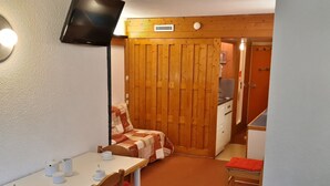 A compact room with a dining table, chairs, a bed with a quilt, a TV mounted on the wall, and a wooden door leading to a kitchenette. - Cozy ski-in/ski-out studio with balcony (Les Arcs 1800)