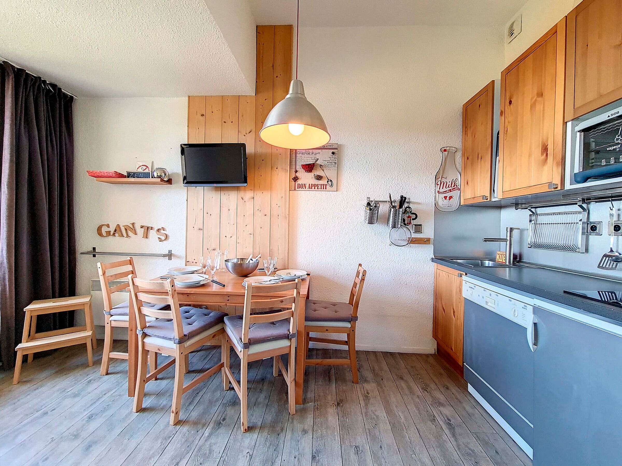 A compact kitchen with wooden cabinets, a dining table with chairs, a hanging light fixture, and a television mounted on the wall.