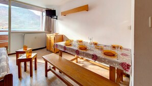 Een eetgedeelte met een tafel voor vier personen, houten banken, een radiator, een raam met uitzicht op bergen en een bevestigde televisie.