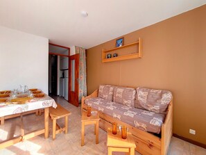 Ein gemütliches Wohnzimmer mit einer Couch, einem Esstisch komplett mit Geschirr und Gläsern, und einer durch eine offene Tür einsehbaren Küchenzeile.
