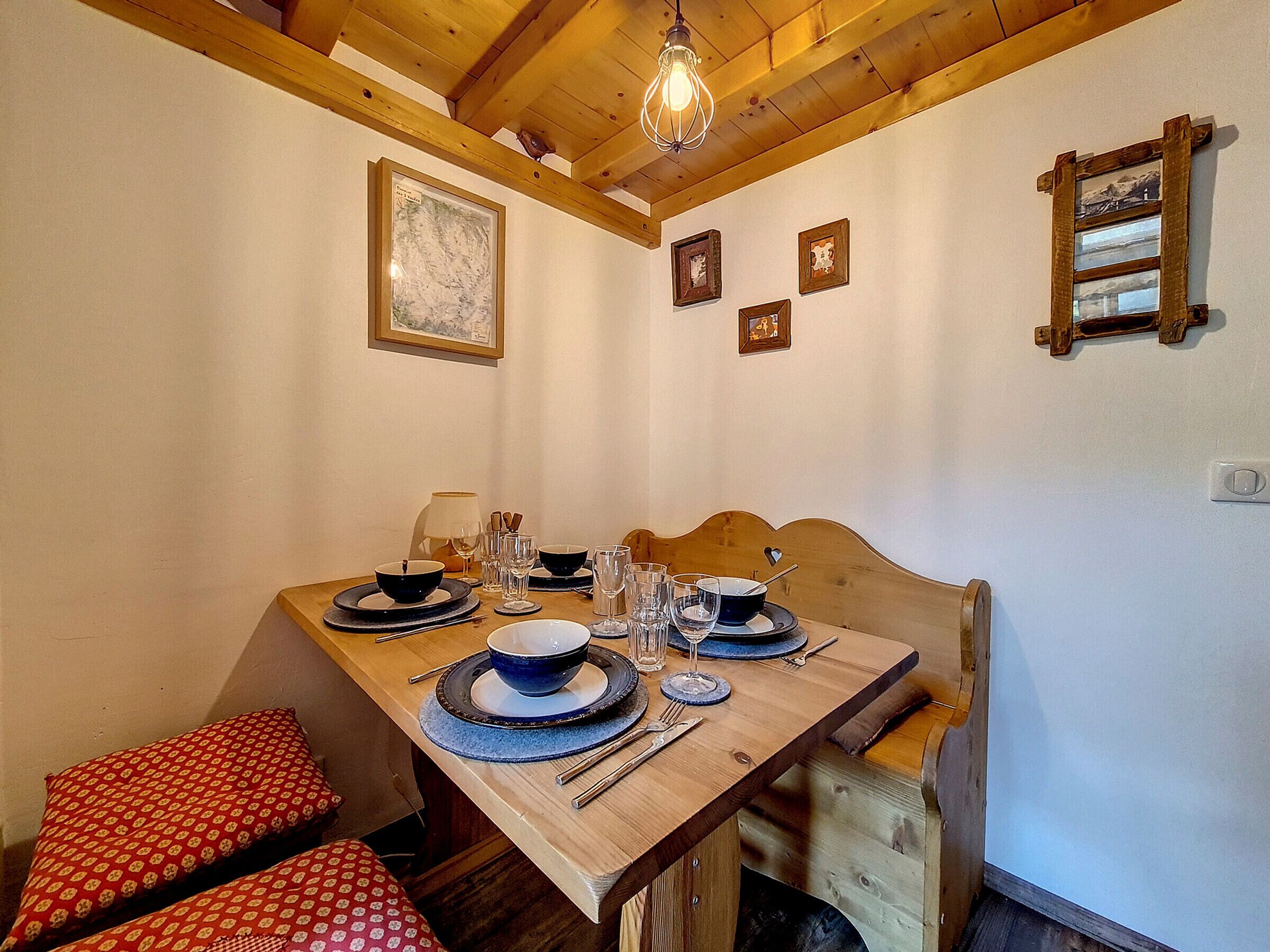 A dining area with a wooden table set for four, including plates, bowls, glasses, and cutlery. There are two benches with red cushions, a wooden ceiling with exposed beams, and framed pictures on the walls.