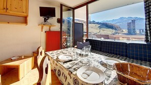 A dining area with a table set for two, a television mounted on the wall, and a view of mountains and buildings through large windows. - Bright studio with balcony on the slopes (Les Menuires)