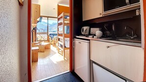 A compact kitchen with a microwave, coffee maker, and sink. A small refrigerator is visible, and the room has a wooden floor and a view of mountains through a large window. - Bright studio with balcony on the slopes (Les Menuires)