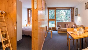 A cozy room with a wooden ladder, a bed, a sofa, a table with cups and a kettle, and large windows offering a view of trees. - 2-Room Apartment · Balcony · Free Wifi (Les Arcs 1800)