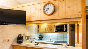 A compact kitchen with wooden cabinetry, a built-in microwave, a coffee maker, and a clock on the wall. - 2-Room Apartment · Balcony · Free Wifi (Les Arcs 1800)