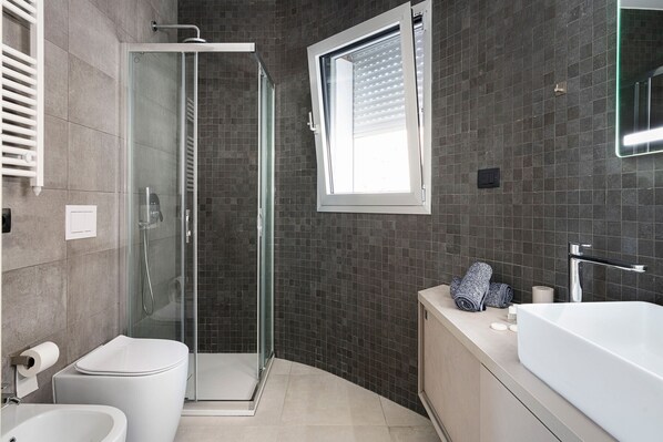 Un baño moderno con ducha envuelta en vidrio, inodoro, lavabo con espejo y ventana.