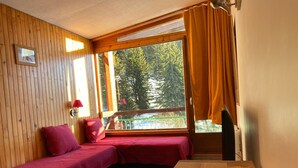 A room with a wooden floor, a bench with a red cover, a window with curtains, and a view of trees. - Cozy Studio · Ski-In/Ski-Out · Balcony · Mountain Views (Les Arcs 1800)