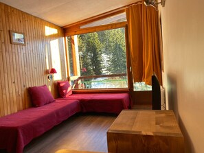 Ein Zimmer mit Holzboden, einer Bank mit roter Sitzfläche, einem Fenster mit Vorhängen und Blick auf Bäume.