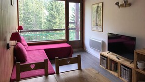 A room with a bed, a table, a TV, and a window with a view of trees. - Cozy Studio · Ski-In/Ski-Out · Balcony · Mountain Views (Les Arcs 1800)