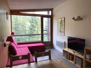 Ein Zimmer mit einem Bett, einem Tisch, einem Fernseher und einem Fenster mit Blick auf Bäume.
