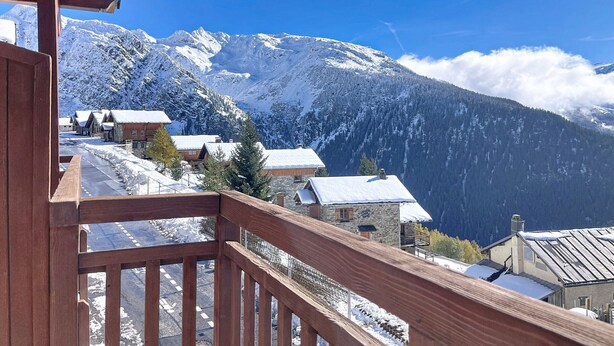 A wooden balcony overlooking a snow-covered mountain village with rustic stone buildings and a clear blue sky.