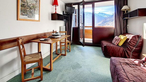 A room with a wooden desk, a red lamp, a brown sofa, and a large window with a mountain view. - Studio at the Foot of the Slopes · South Balcony (Les Menuires)
