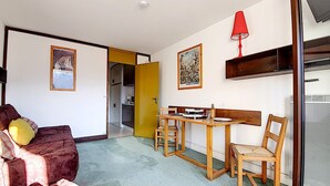 A compact living space with a sofa, a dining table with chairs, a red lamp, and a kitchen area visible through an open door. - Studio at the Foot of the Slopes · South Balcony (Les Menuires)