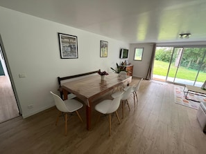 A dining area with a wooden table and four chairs, a small cabinet with a TV, and a view of a green landscape through large windows. - Charming 2BR Garden Apt with Pool Access near Basque Coast & Bayonne (Urt)