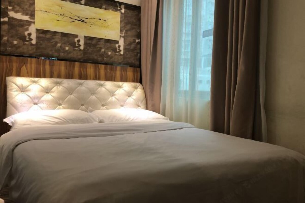 A neatly made bed with a tufted headboard, a plain wall with a piece of abstract art, and a window with curtains.