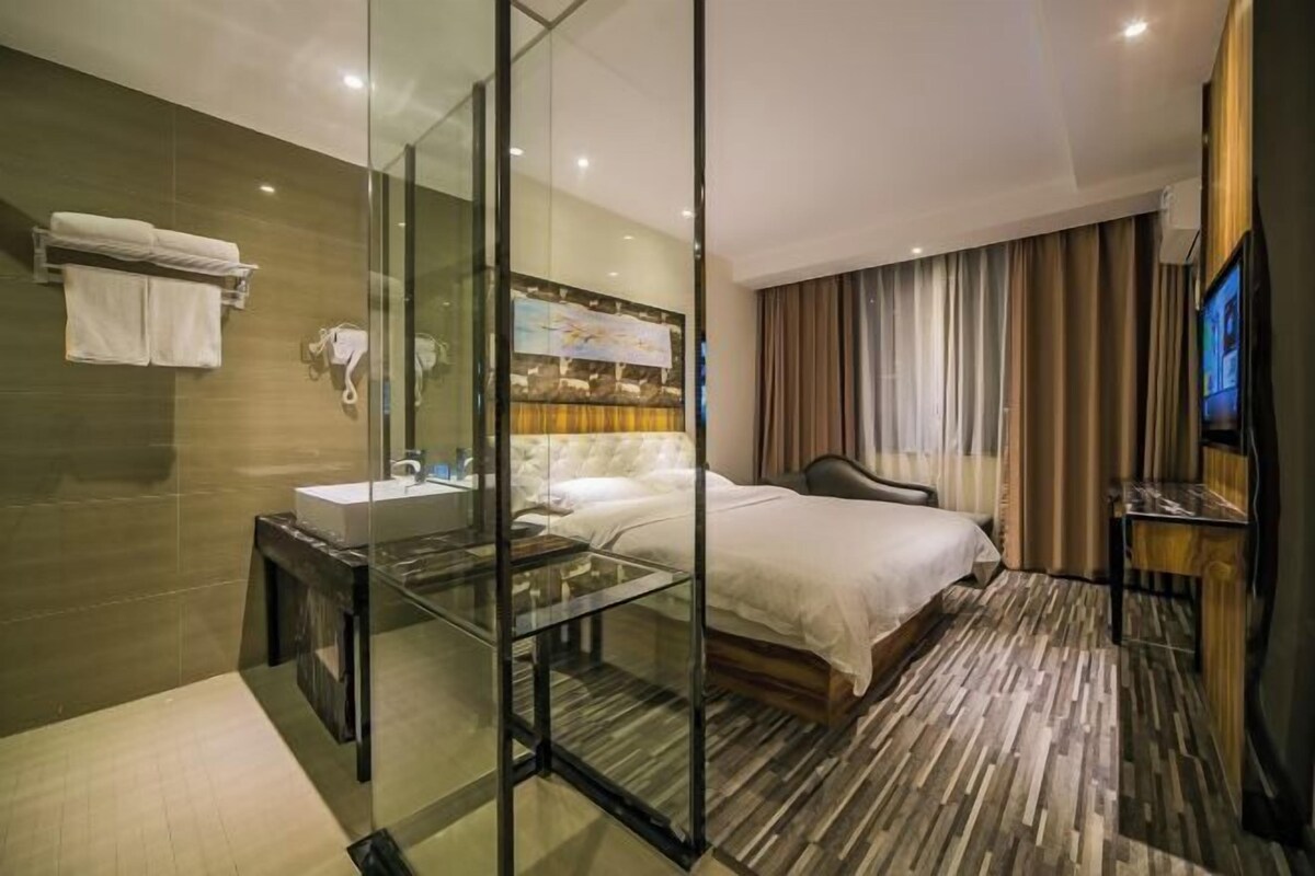 A modern hotel room with a glass-enclosed shower, a large bed, a flat-screen TV, and a wooden floor.
