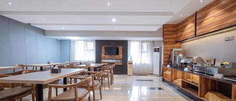A spacious dining area with multiple tables and chairs, a kitchen counter with appliances, and a large screen on the wall.