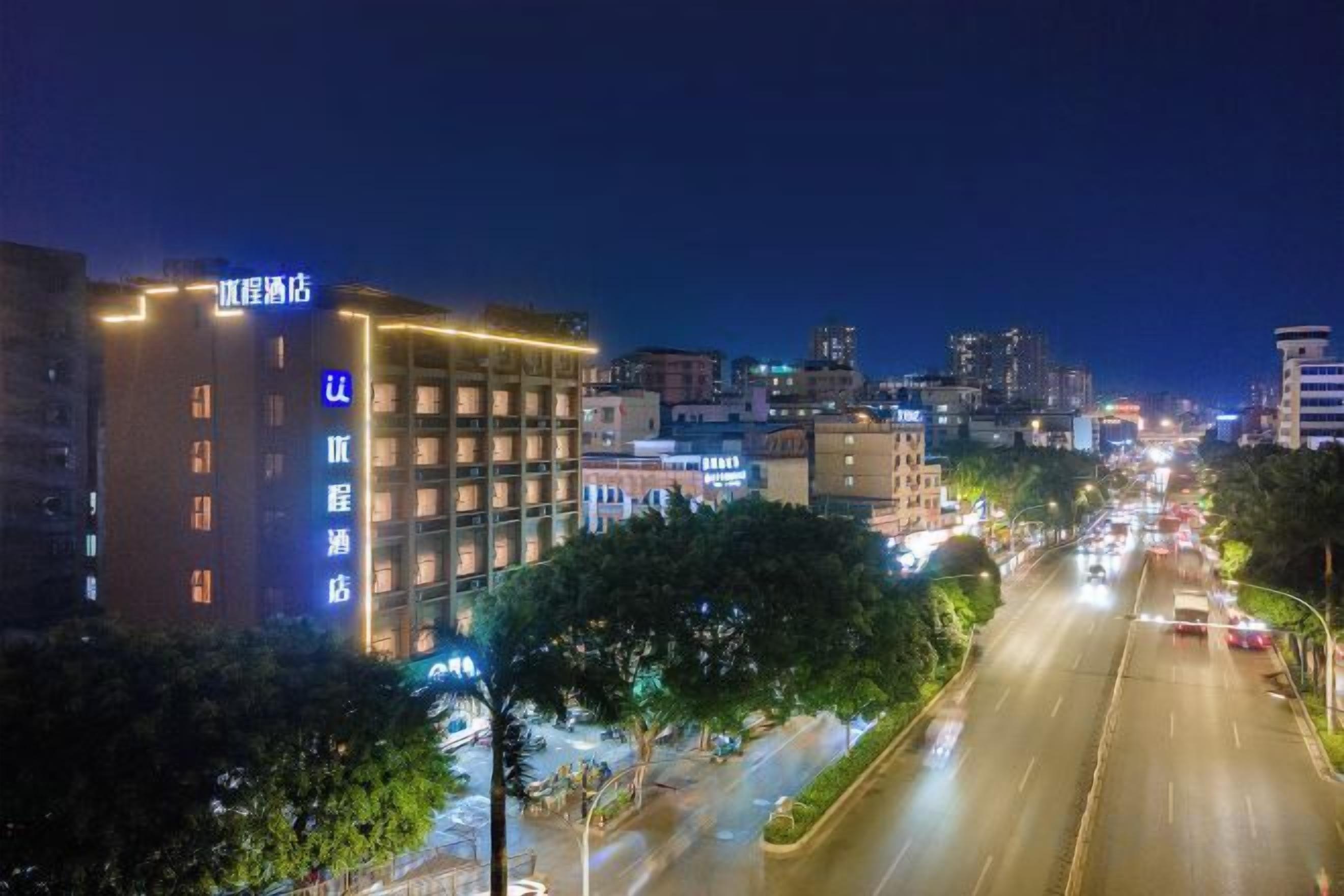 Un paisaje urbano nocturno con edificios iluminados, un hotel de varios pisos con un cartel, y una calle concurrida con vehículos.