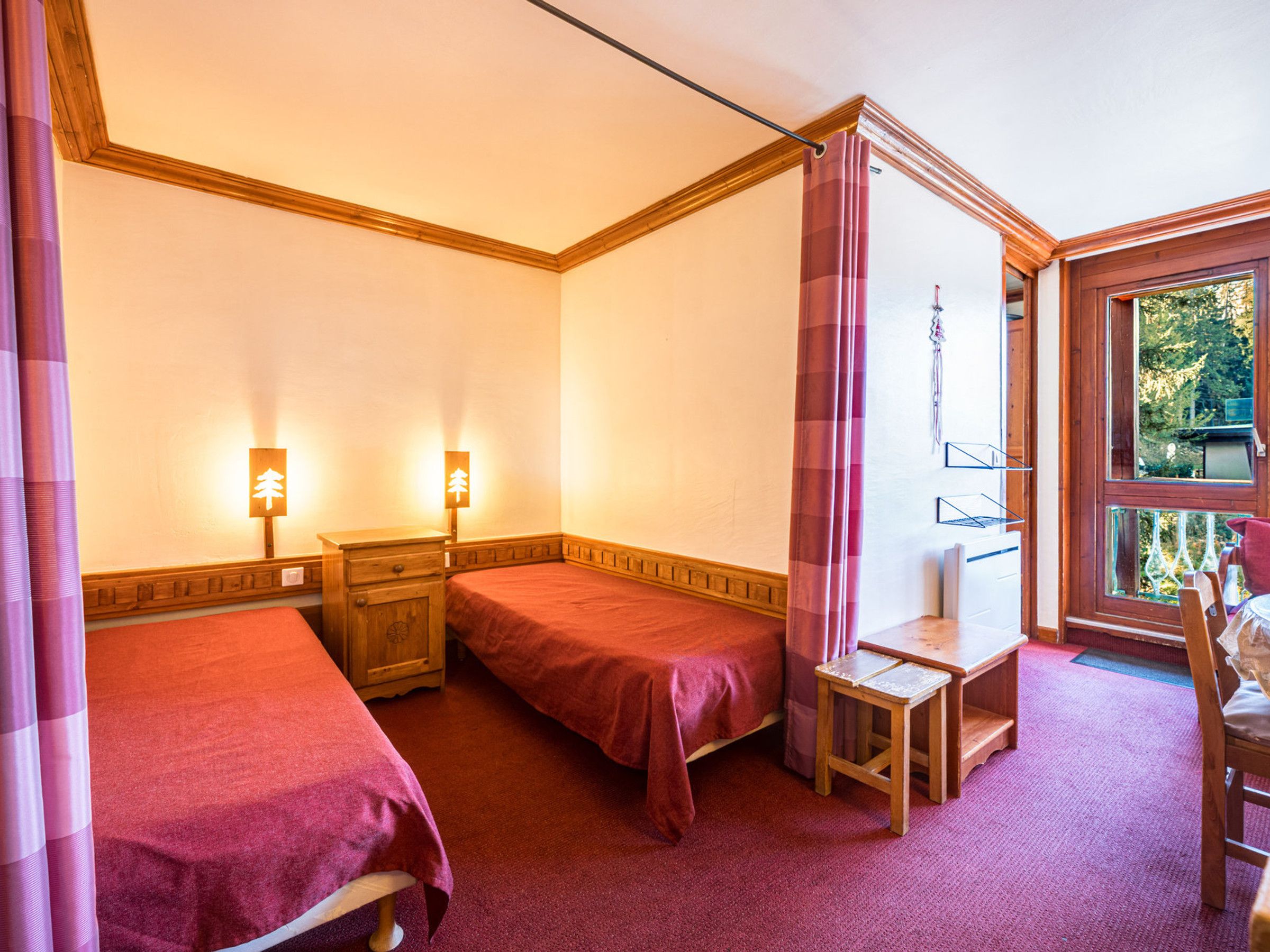 A room with two beds, a wooden nightstand, a small table, and a window with curtains.