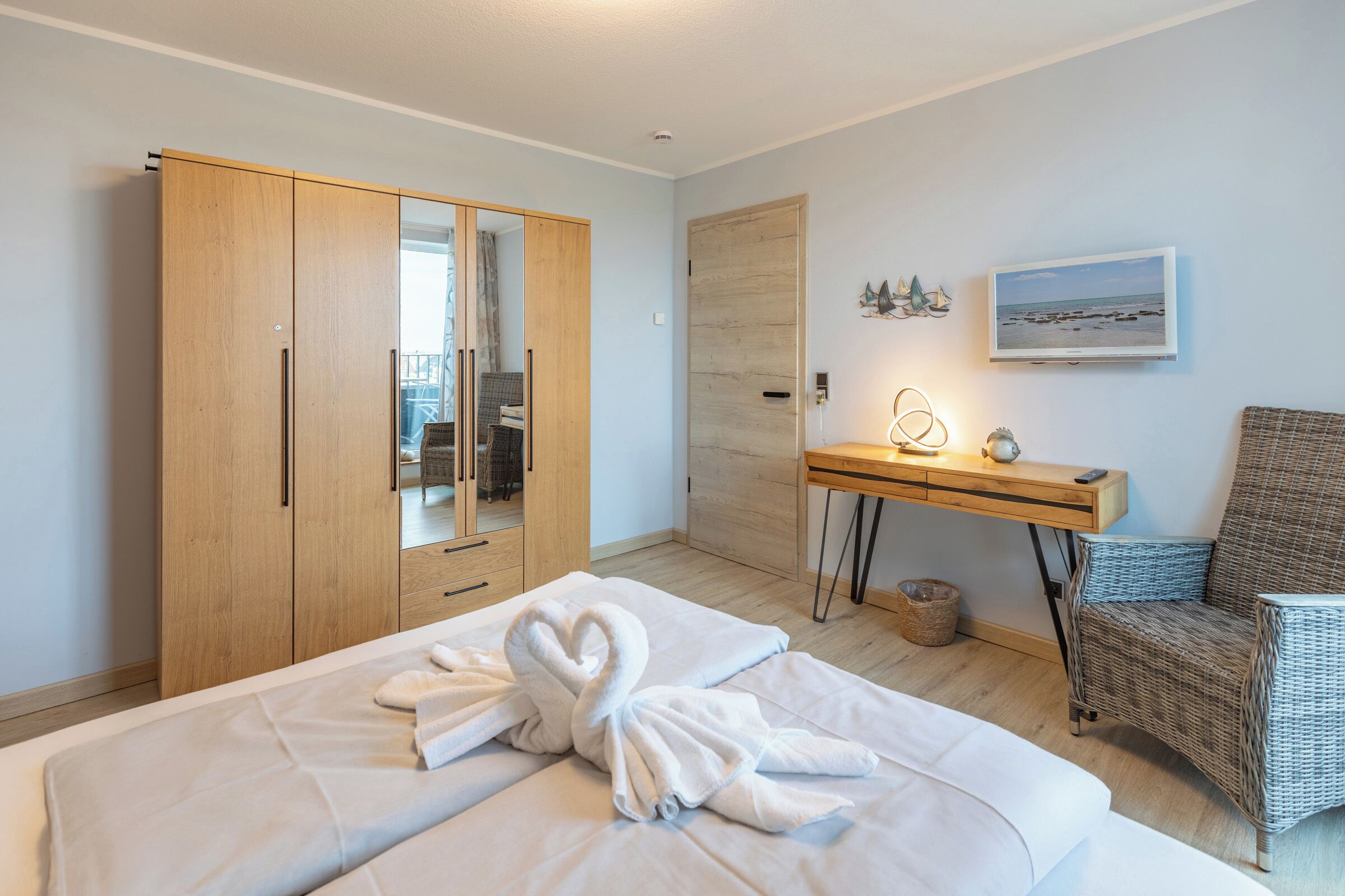 A hotel room with a bed, a wooden wardrobe, a desk with a lamp, a wicker chair, and a beach view painting.