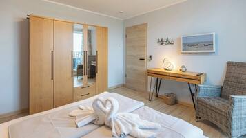 A hotel room with a bed, a wooden wardrobe, a desk with a lamp, a wicker chair, and a beach view painting.