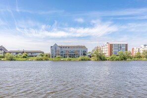 A riverside view of modern buildings with flat roofs and large windows, surrounded by greenery.