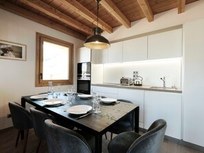 Un comedor con una mesa de madera dispuesta para cuatro, una lámpara colgante y vista a la cocina a través de un ventanal abierto.