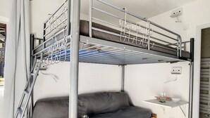 Kerrossängyssä on harmaa patja ja alapuolella puinen alusta. Oikealla puolella on pieni hylly, jonka päällä on kulho ja laseja.