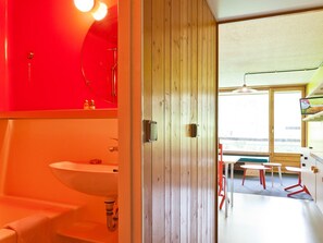 Ein modernes Badezimmer mit roter Akzentwand, einem Waschbecken und einer Holztür, die in einen Essbereich mit roten Stühlen und einem Tisch führt.