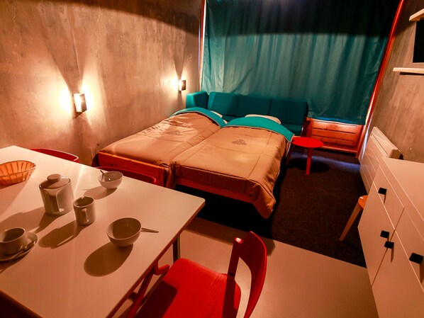 A compact room with a bed, a table with cups and a bowl, a chair, and a small table with a red chair.