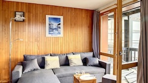 Ein modernes Wohnzimmer mit einem Ecksofa, einem kleinen Holztisch mit Teller und Gläsern sowie einem großen Fenster mit Blick ins Freie.