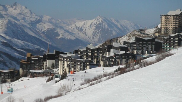A ski resort with modern buildings, a ski slope, and snow-covered mountains in the background.