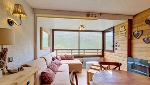 A cozy room with a wooden table, a sofa with patterned cushions, a window with a view of the outdoors, and a wall-mounted lamp.