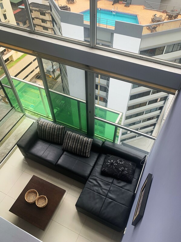 A modern living room with a black leather sofa, two cushions, and a small wooden table with two woven bowls. A large window offers a view of a swimming pool and outdoor seating area. - Modern loft-style, 2 level apartment  with city views, cover parking and pool.   (Panamá)